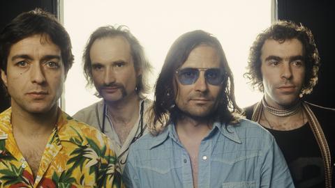 Frontales Porträt der Rockband Can vor einem weiß überstrahlte Fenster: Jaki Liebezeit, Holger Czukay, Irmin Schmidt und Peter Gilmour stehen nebeneinander. In der von gedeckten Farben dominierten Kleidung sticht Liebezeit farbenfrohes Hawaiihemd heraus.