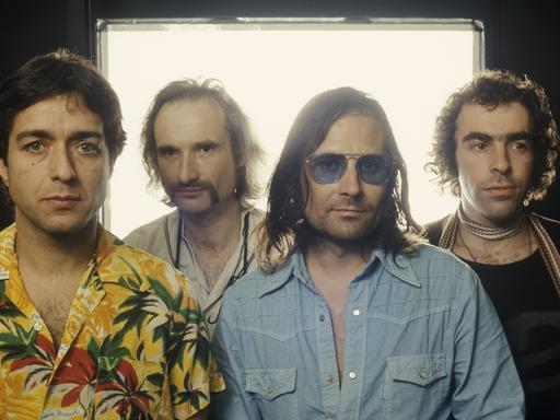Frontales Porträt der Rockband Can vor einem weiß überstrahlte Fenster: Jaki Liebezeit, Holger Czukay, Irmin Schmidt und Peter Gilmour stehen nebeneinander. In der von gedeckten Farben dominierten Kleidung sticht Liebezeits farbenfrohes Hawaiihemd heraus.