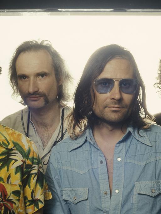 Frontales Porträt der Rockband Can vor einem weiß überstrahlte Fenster: Jaki Liebezeit, Holger Czukay, Irmin Schmidt und Peter Gilmour stehen nebeneinander. In der von gedeckten Farben dominierten Kleidung sticht Liebezeits farbenfrohes Hawaiihemd heraus.