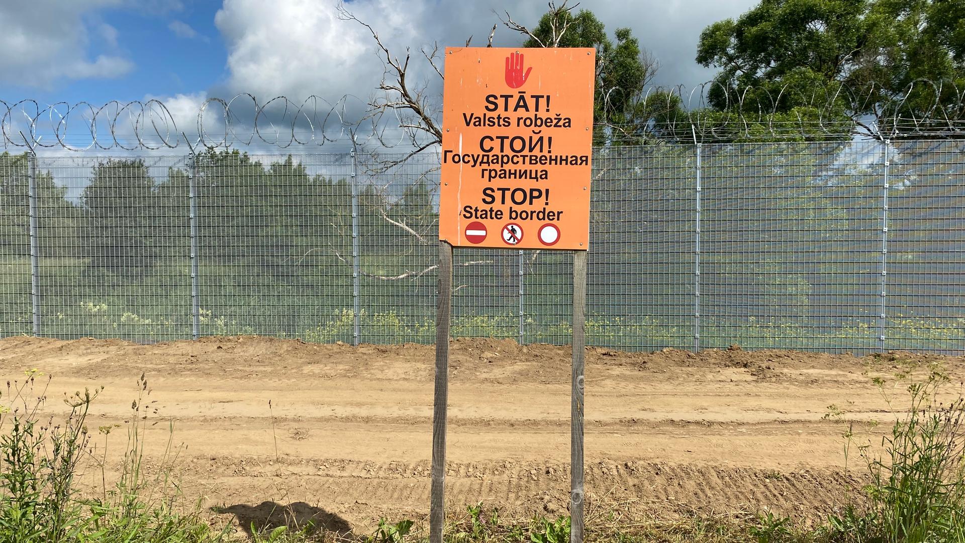 "Halt!- Staatsgrenze" steht in drei Sprachen auf einem Schild an der lettischen Grenze zu Russland. Im Hintergrund ist ein Zaun zu sehen, der mit Stacheldraht gesichert ist.