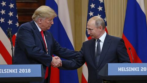 Donald Trump und Wladimir Putin beim Gipfeltreffen in Helsinki 2018. Sie gehen aufeinander zu und geben sich die Hand. Beide schauen sich an und gucken freundlich. Im Hintergrund die Fahnen beider Länder. 