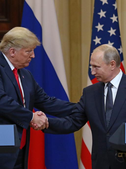 Donald Trump und Wladimir Putin beim Gipfeltreffen in Helsinki 2018. Sie gehen aufeinander zu und geben sich die Hand. Beide schauen sich an und gucken freundlich. Im Hintergrund die Fahnen beider Länder. 