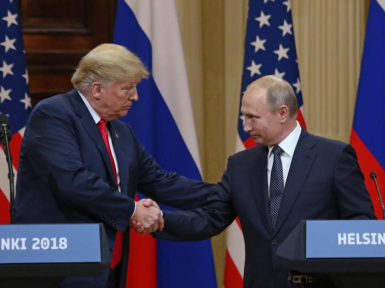 Donald Trump und Wladimir Putin beim Gipfeltreffen in Helsinki 2018. Sie gehen aufeinander zu und geben sich die Hand. Beide schauen sich an und gucken freundlich. Im Hintergrund die Fahnen beider Länder. 