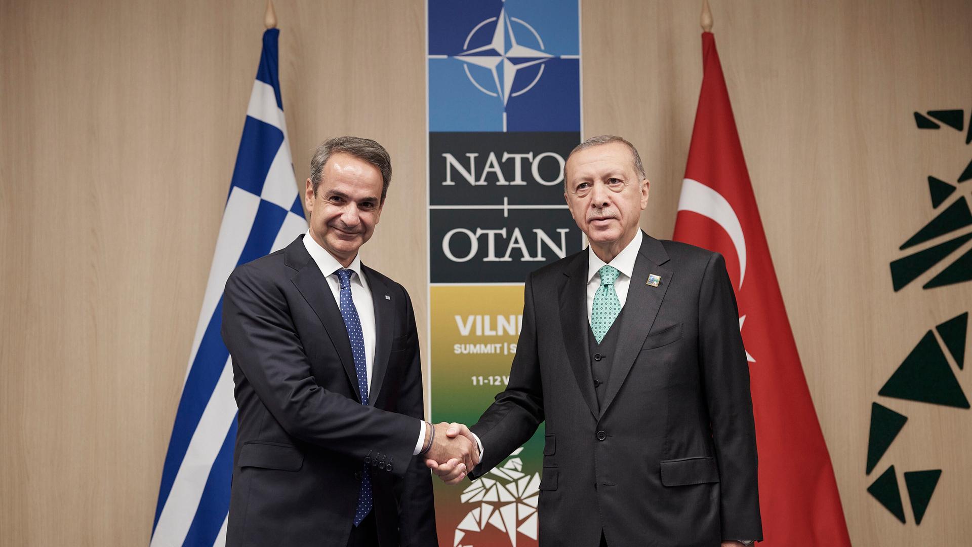 Zwei Männer schütteln sich die Hände und schauen in die Kamera, im Hintergrund sind Fahnen von Griechenland und der Türkei zu sehen sowie ein Log der NATO. Zwei Männer schütteln sich die Hände und schauen in die Kamera, im Hintergrund sind Fahnen von Griechenland und der Türkei zu sehen sowie ein Log der NATO.