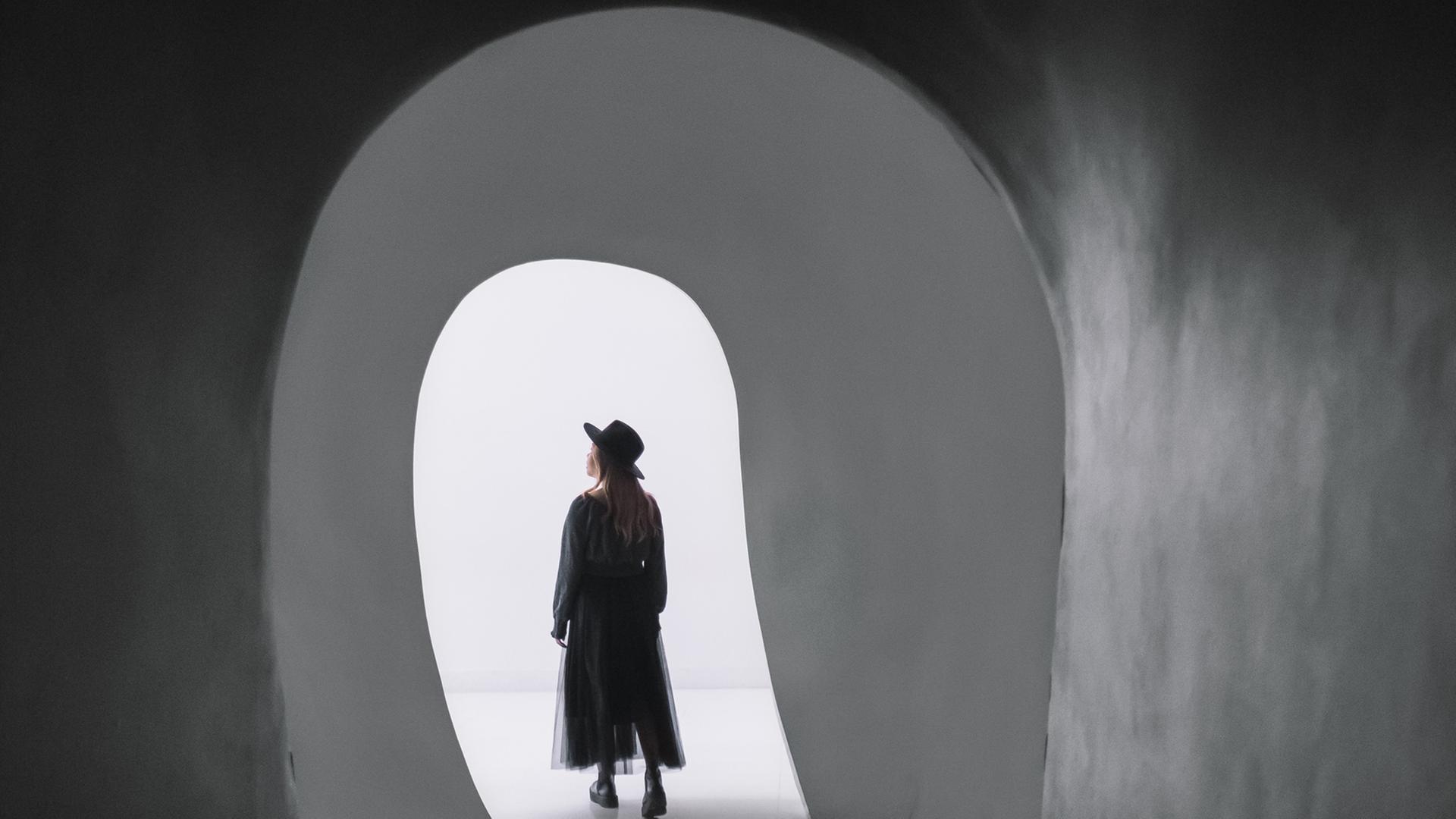Silke macht sich auf den Weg, Annas Geheimnis zu lüften. Zu sehen: Eine junge Frau mit Schlapphut, kommt aus einem schwarzen Tunnel und geht in die Helligkeit hinein.