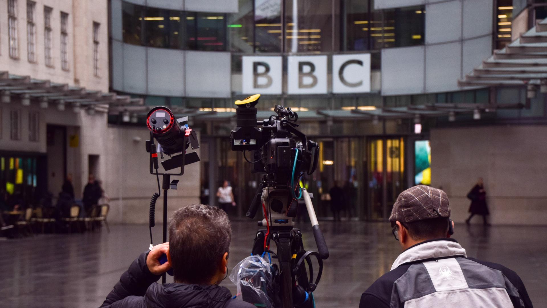 Blick auf das BBC-Gebäude am Tag der Pressekonferenz, während der die BBC-Führung zurücktrat. Zu sehen ist der BBC-Schriftzug an einer verglasten Gebäudehülle, im Vordergrund Mikros und Kameras. 