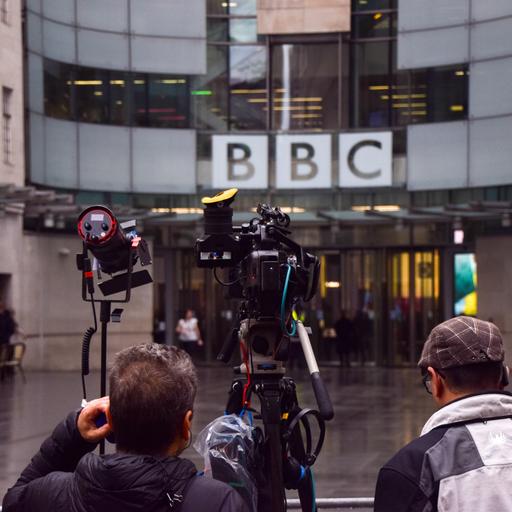 Blick auf das BBC-Gebäude am Tag der Pressekonferenz, während der die BBC-Führung zurücktrat. Zu sehen ist der BBC-Schriftzug an einer verglasten Gebäudehülle, im Vordergrund Mikros und Kameras. 