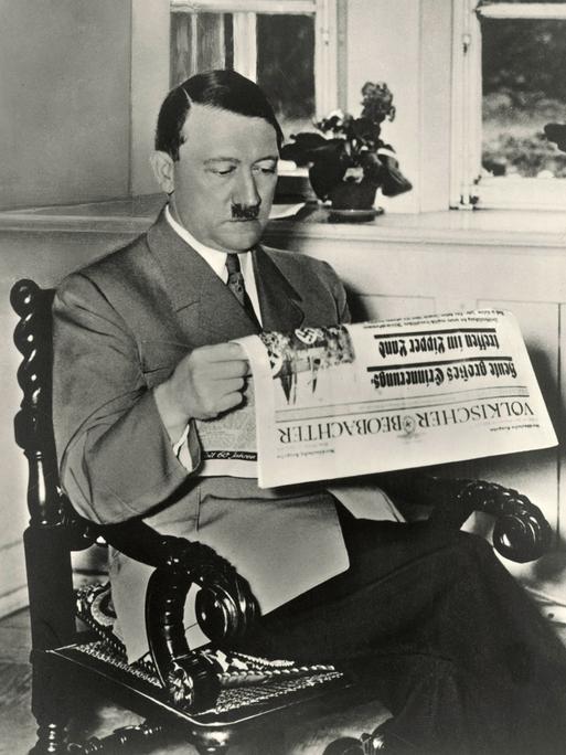 Adolf Hitler sitzt in einem Sessel und schaut auf die Titelseite der Zeitung "Völkischer Beobachter".