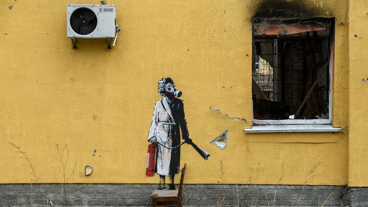 Ein Graffitti von dem Künstler Banksy an der Wand eines zerstörten gelben Wohnhauses in einem Vorort von Kiew in der Ukraine. Es zeigt eine Frau im Bademantel mit einer Gasmaske, die mit einem Feuerlöscher in der Hand neben einem zerstörtem Fenster steht.