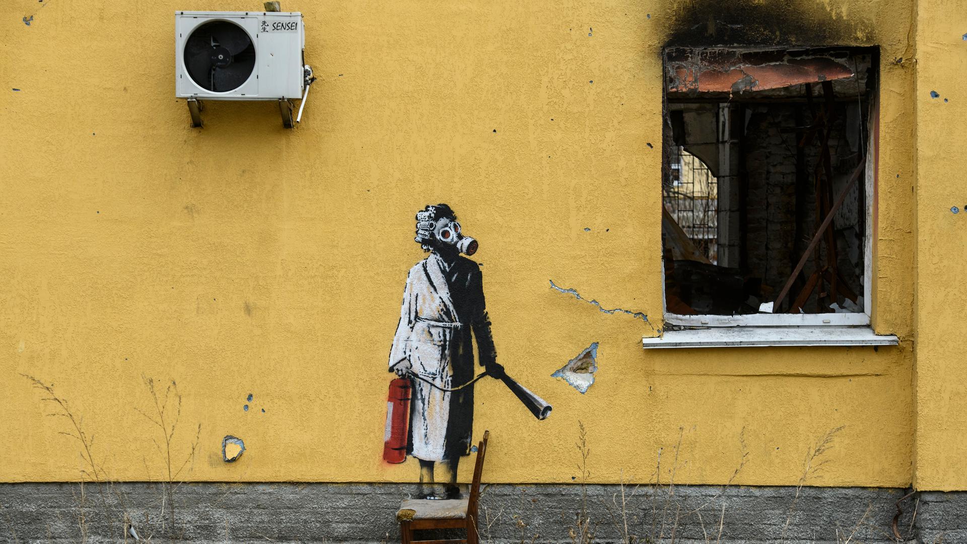 Ein Graffitti von dem Künstler Banksy an der Wand eines zerstörten gelben Wohnhauses in einem Vorort von Kiew in der Ukraine. Es zeigt eine Frau im Bademantel mit einer Gasmaske, die mit einem Feuerlöscher in der Hand neben einem zerstörtem Fenster steht.