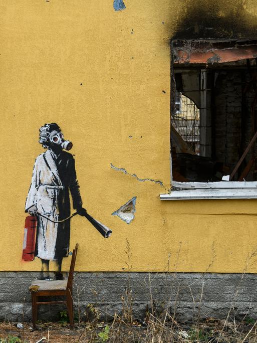 Ein Graffitti von dem Künstler Banksy an der Wand eines zerstörten gelben Wohnhauses in einem Vorort von Kiew in der Ukraine. Es zeigt eine Frau im Bademantel mit einer Gasmaske, die mit einem Feuerlöscher in der Hand neben einem zerstörtem Fenster steht. Ein Graffitti von dem Künstler Banksy an der Wand eines zerstörten gelben Wohnhauses in einem Vorort von Kiew in der Ukraine. Es zeigt eine Frau im Bademantel mit einer Gasmaske, die mit einem Feuerlöscher in der Hand neben einem zerstörtem Fenster steht.