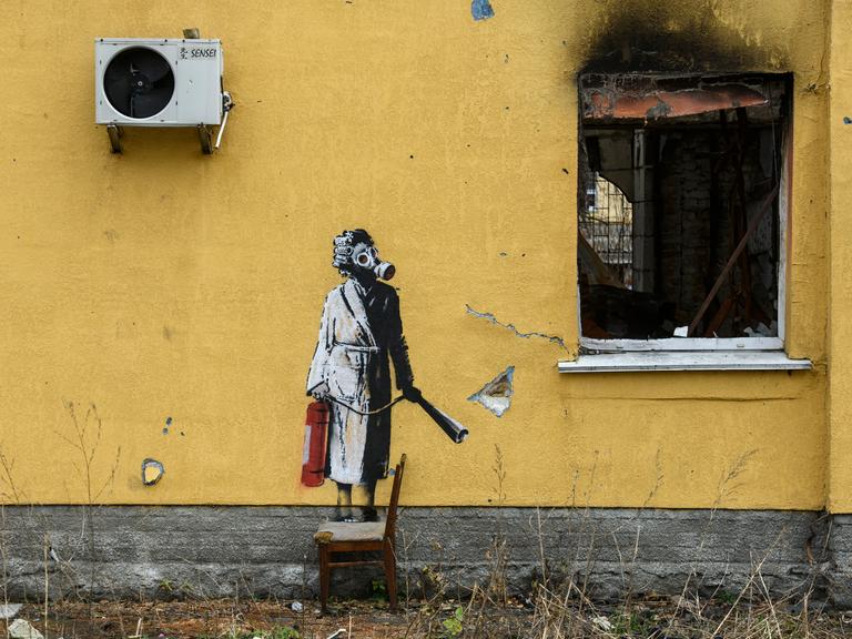 Ein Graffitti von dem Künstler Banksy an der Wand eines zerstörten gelben Wohnhauses in einem Vorort von Kiew in der Ukraine. Es zeigt eine Frau im Bademantel mit einer Gasmaske, die mit einem Feuerlöscher in der Hand neben einem zerstörtem Fenster steht. Ein Graffitti von dem Künstler Banksy an der Wand eines zerstörten gelben Wohnhauses in einem Vorort von Kiew in der Ukraine. Es zeigt eine Frau im Bademantel mit einer Gasmaske, die mit einem Feuerlöscher in der Hand neben einem zerstörtem Fenster steht.