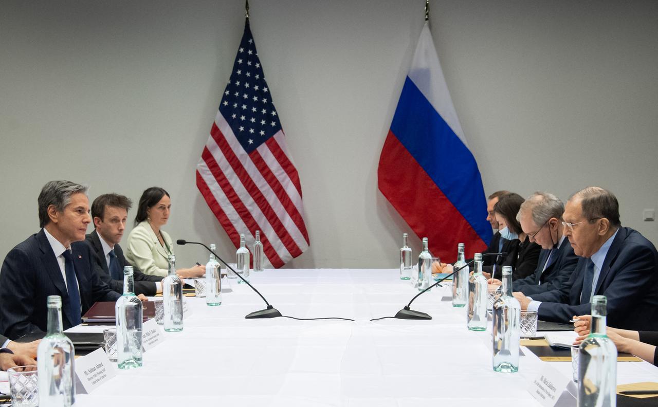 Antony Blinken und Sergej Lawrow sitzen mit Mitarbeitern an einem großen Tisch. Im Hintergrund stehen eine amerikanische und eine russische Fahne. 