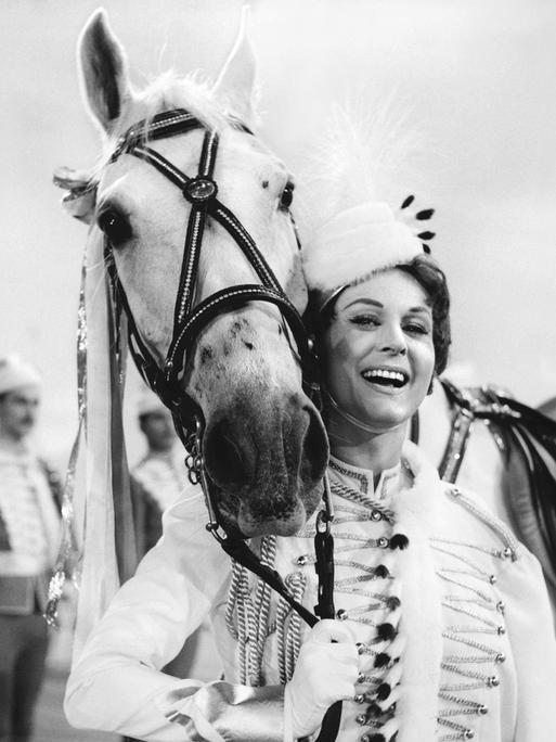 Marika Rökk trägt eine ungarische Tracht und lehnt lachend an einem Pferd. (Filmstill)