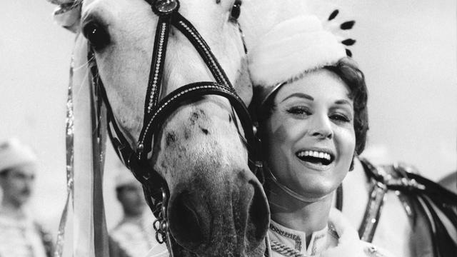 Marika Rökk trägt eine ungarische Tracht und lehnt lachend an einem Pferd. (Filmstill)