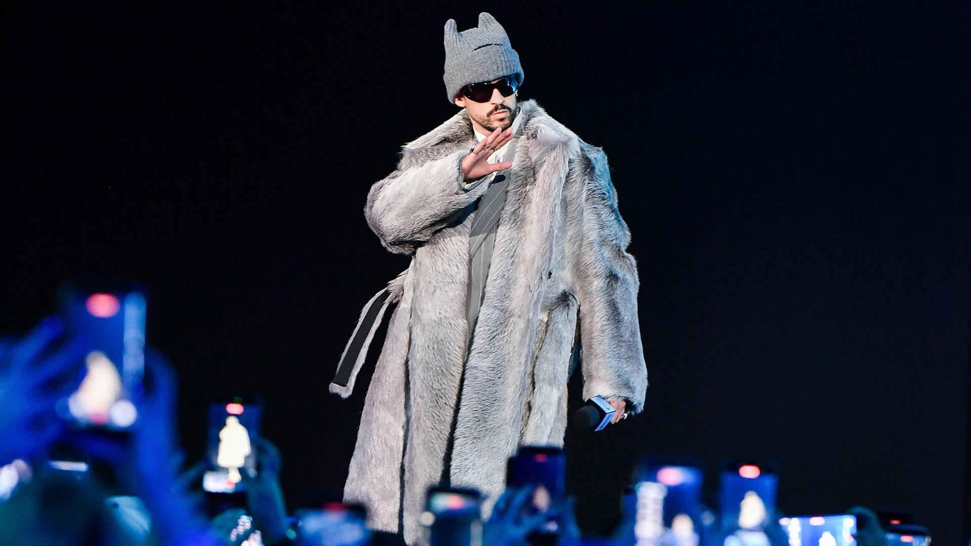 Der puertoricanische Musiker steht auf einer Bühne unter Scheinwerfern vor einem Publikum, trägt ein langes, helles Fell-Outfit und eine Mütze.