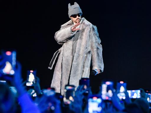 Der puertoricanische Musiker steht auf einer Bühne unter Scheinwerfern vor einem Publikum, trägt ein langes, helles Fell-Outfit und eine Mütze.