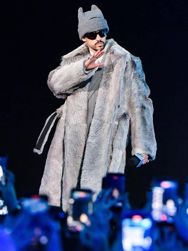 Der puertoricanische Musiker steht auf einer Bühne unter Scheinwerfern vor einem Publikum, trägt ein langes, helles Fell-Outfit und eine Mütze.