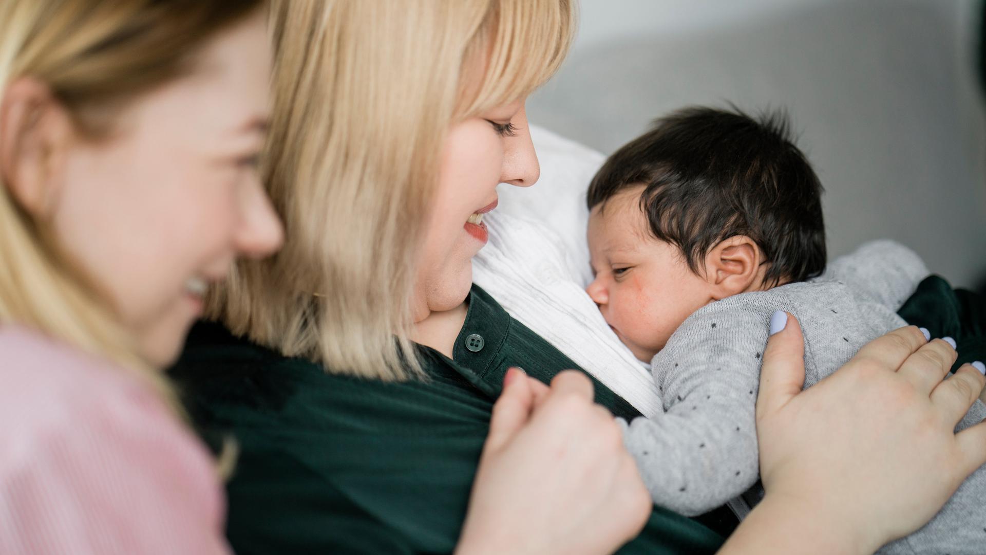 Zwei junge Frauen sitzen auf der Couch, eine hält ein kleines Baby auf dem Arm welches friedlich schläft. Zwei junge Frauen sitzen auf der Couch, eine hält ein kleines Baby auf dem Arm welches friedlich schläft.