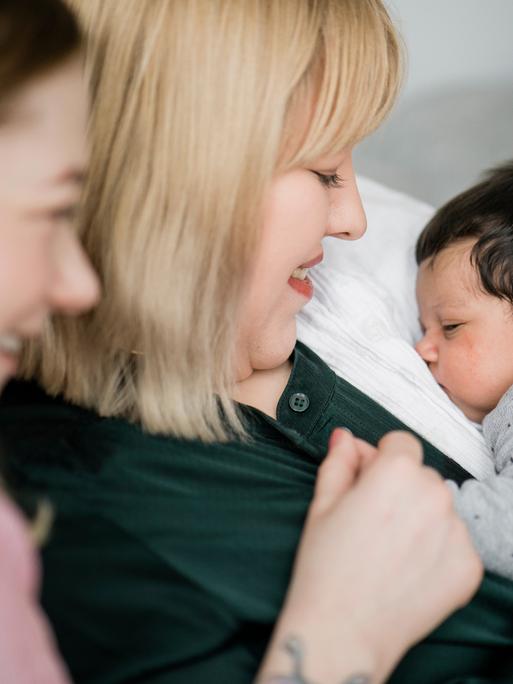 Zwei junge Frauen sitzen auf der Couch, eine hält ein kleines Baby auf dem Arm welches friedlich schläft.