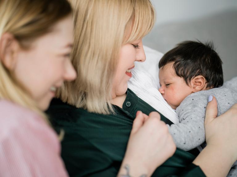 Zwei junge Frauen sitzen auf der Couch, eine hält ein kleines Baby auf dem Arm welches friedlich schläft.