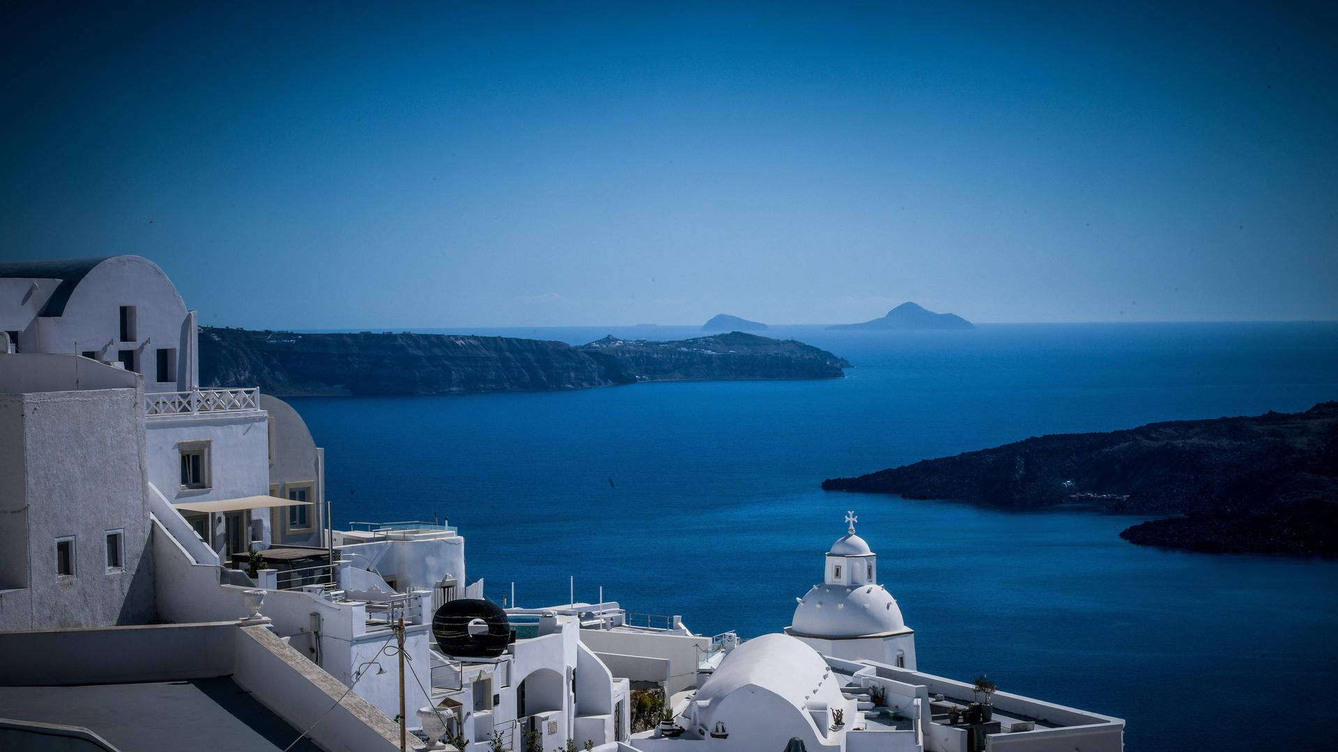 Weiße Häuser auf der griechischen Insel Santorini, im Hintergrund das Mittelmeer und weitere Inseln.