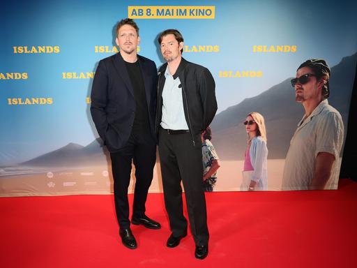 Premiere des Spielfilms "Islands" von Regisseur Jan-Ole Gerster mit Hauptdarsteller Sam Riley. Sie stehen vor einer Kinoleinwand, auf die ein Standbild des Films projiziert ist.