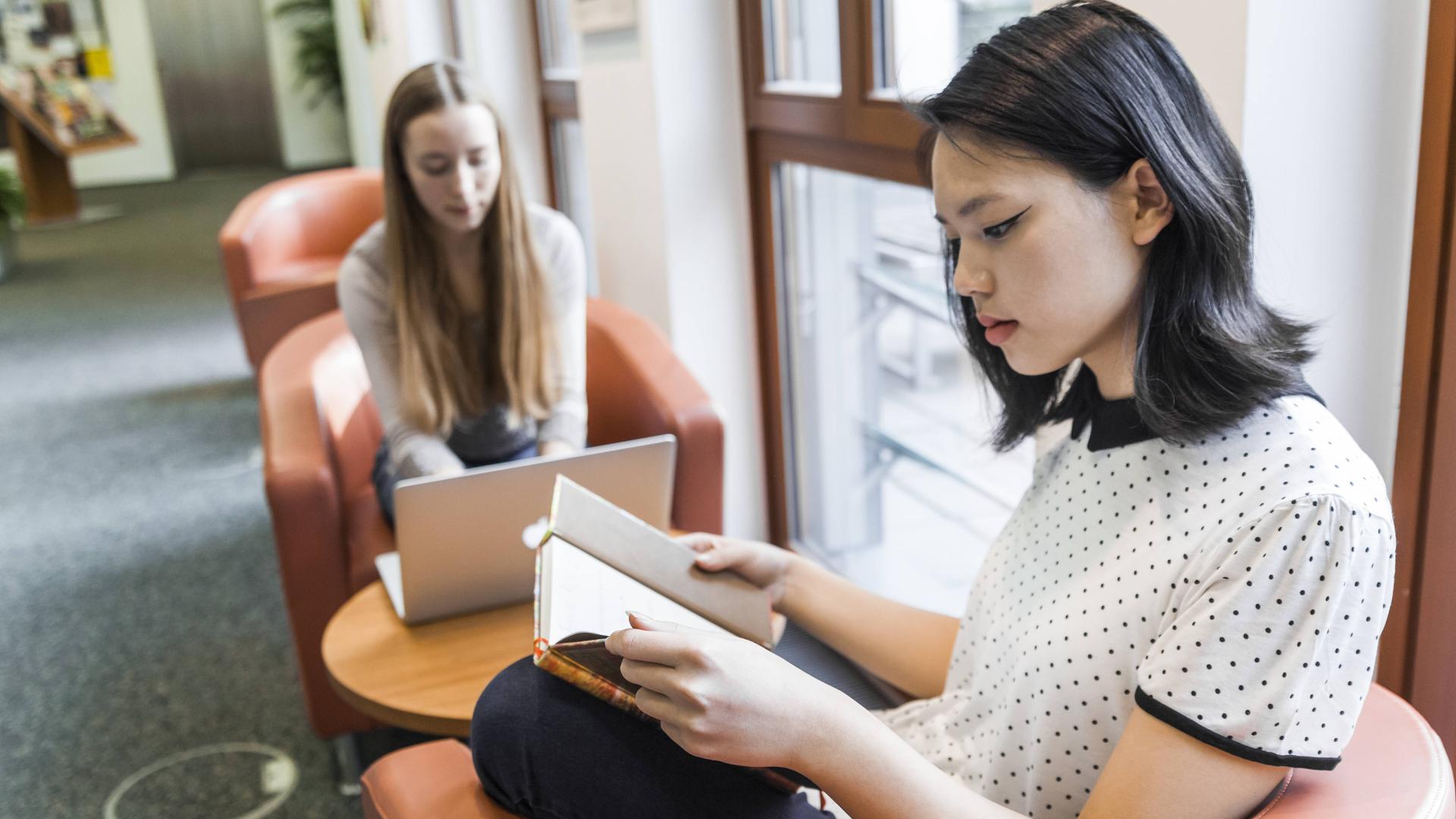 Zwei Mädchen sitzen in Sesseln. Eine schaut in ein Laptop, die andere liest ein Buch. Zwei Mädchen sitzen in Sesseln. Eine schaut in ein Laptop, die andere liest ein Buch.