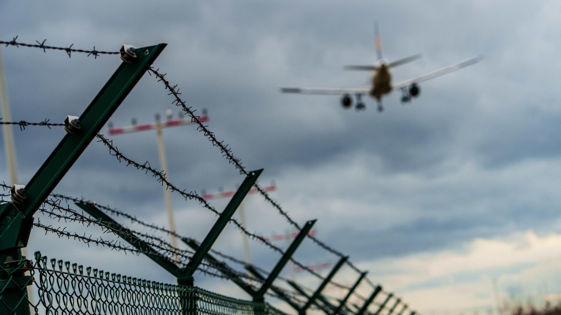 In der unteren Bildhälfte verläuft ein Stacheldraht am Gelände des Rhein-Main-Flughafens in Frankfurt. In der oberen Bildhälfte ist von hinten ein fliegendes Flugzeug zu sehen. In der unteren Bildhälfte verläuft ein Stacheldraht am Gelände des Rhein-Main-Flughafens in Frankfurt. In der oberen Bildhälfte ist von hinten ein fliegendes Flugzeug zu sehen.