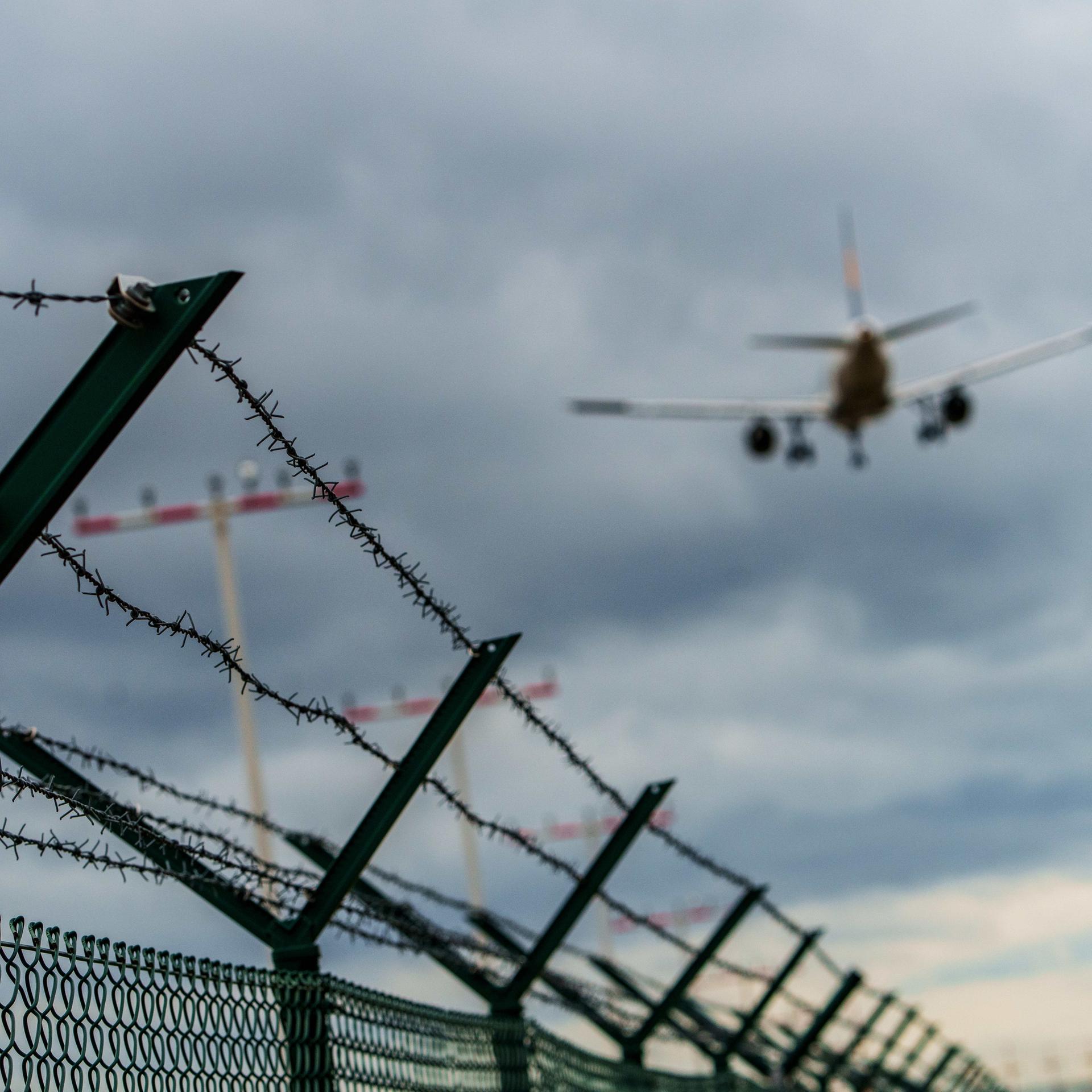 In der unteren Bildhälfte verläuft ein Stacheldraht am Gelände des Rhein-Main-Flughafens in Frankfurt. In der oberen Bildhälfte ist von hinten ein fliegendes Flugzeug zu sehen. 