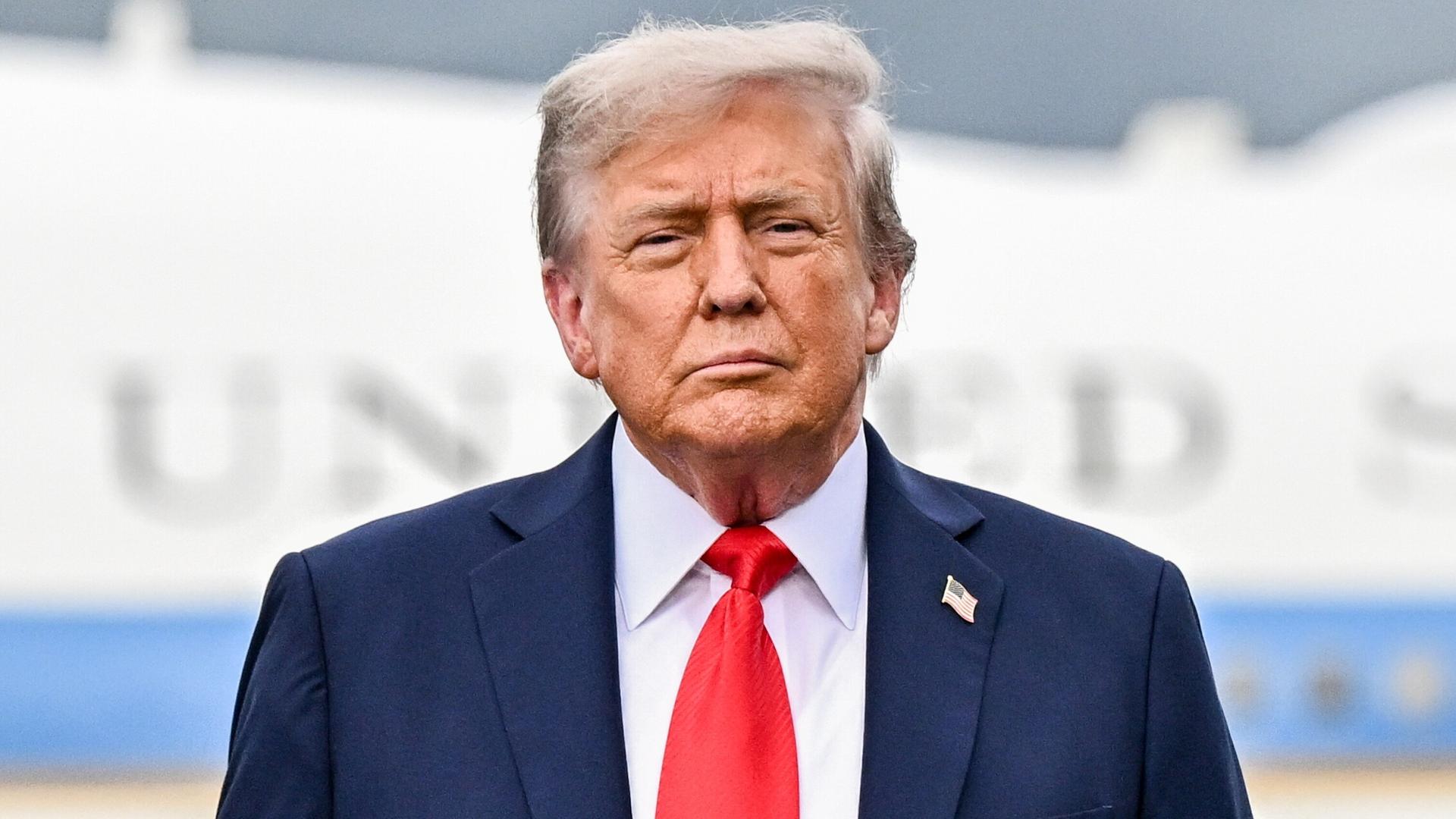 Trump mit schwarzem Jackett und rotzer Krawatte vor seiner Regierungsmaschine, die man nur unscharf sieht. 