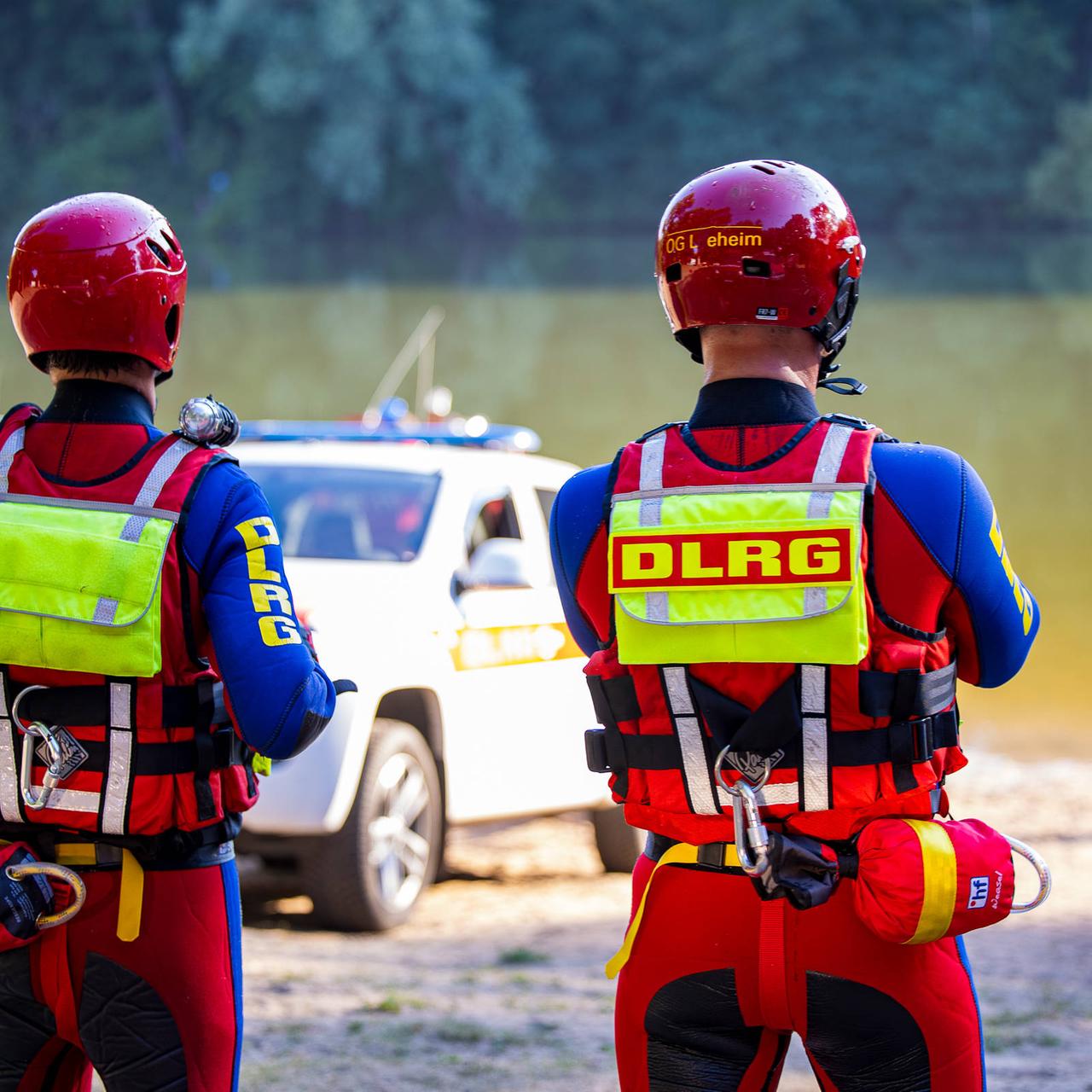Zwei Mitarbeiter von der  DLRG stehen am Ufer von einem See. Sie beobachten Rettungsarbeiten. 