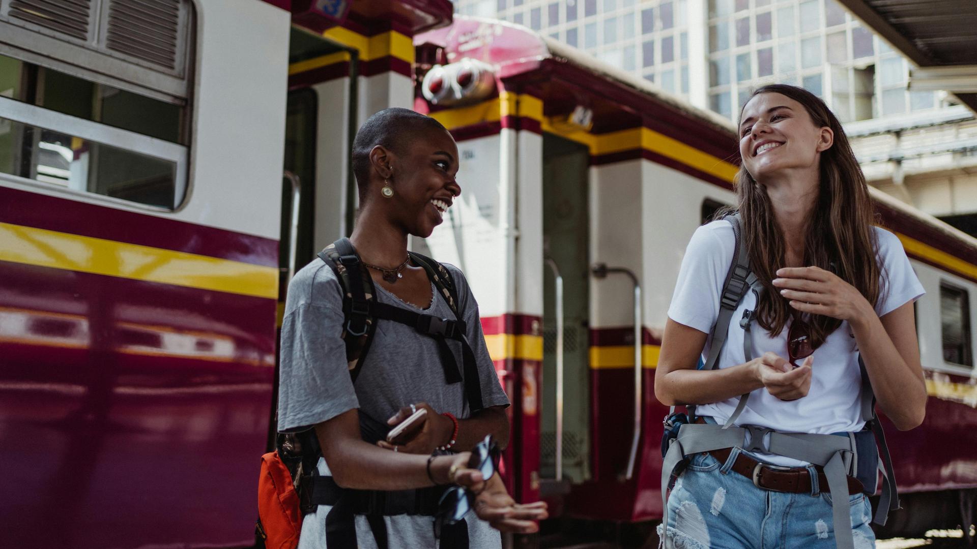 Zwei junge Frauen lachen auf einem Bahnsteigt sich an. Man sieht, dass sie sie gut verstehen.