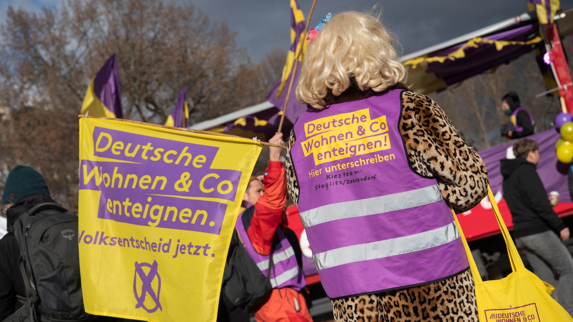 Mit einer Fahne und einer Weste des Bündnisses "Deutsche Wohnen und Co enteignen" steht ein Teilnehmer der Demonstration "Karneval der Enteignung" in Kreuzberg auf dem Oranienplatz. Er trägt eine blonde Perücke. Um ihn herum werden Transparente hochgehalten.