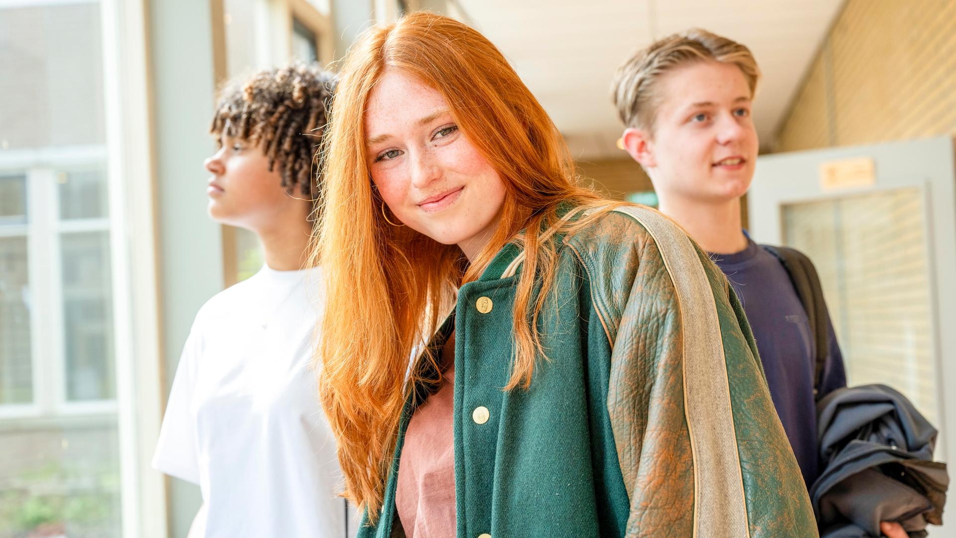 Eine lächelnde junge rothaarige Frau mit zwei männlichen Teenagern im Hintergrund steht in einem Schulkorridor. 