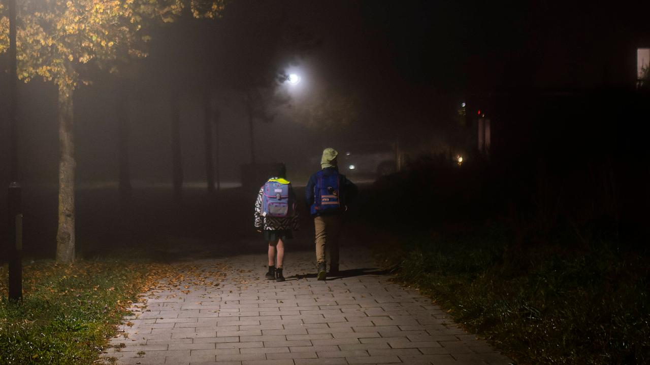 Zwei Kinder mit Schulranzen gehen bei Dunkelheit und Nebel einen Weg entlang, der von Straßenlaternen beleuchtet wird. 