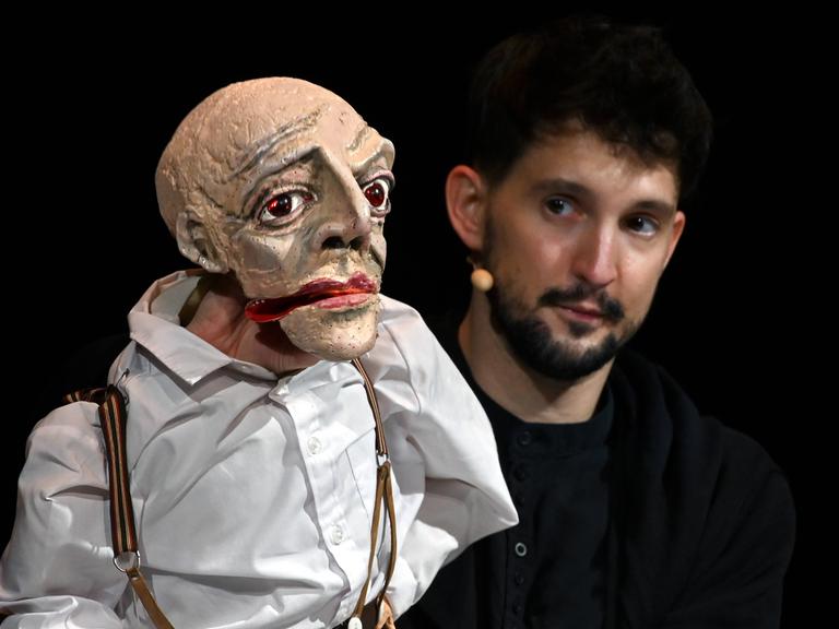 Der Puppenspieler Nikolaus Habjan mit einer Puppe bei einem Auftritt vor schwarzem Hintergrund.
