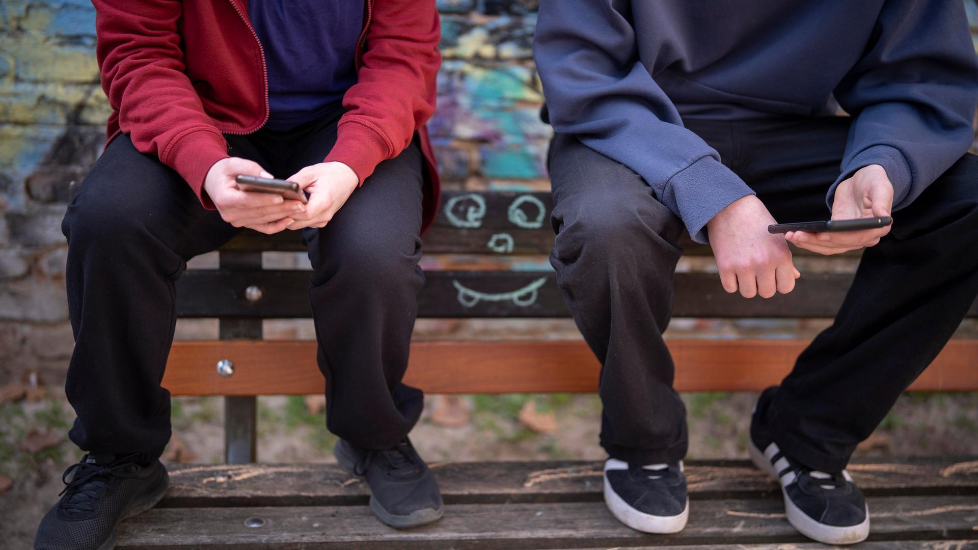 Jugendliche sitzen vor einer Graffiti-Wand auf einer Bank und schauen auf ihre Handys.