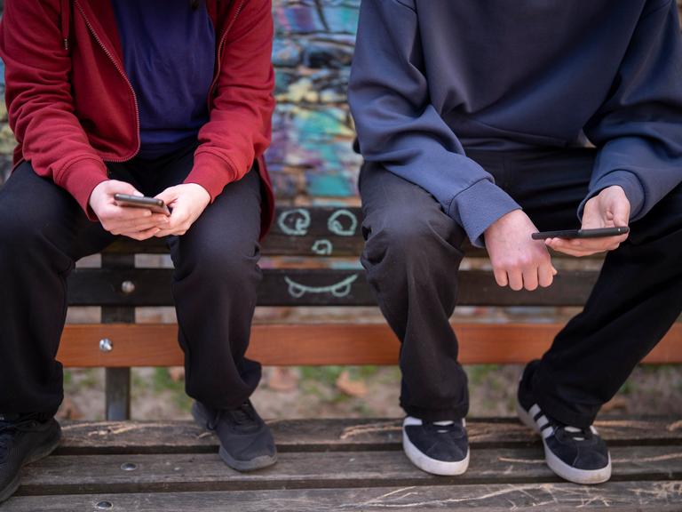Jugendliche sitzen vor einer Graffiti-Wand auf einer Bank und schauen auf ihre Handys.
