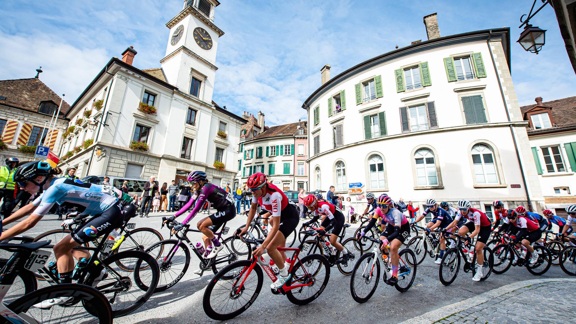 Zahlreiche Radsportlerinnen fahren dicht hintereinander durch das Zentrum einer teilnehmenden Stadt. 