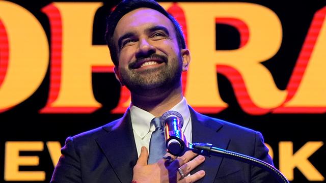 Zohran Mamdani lacht und hält sich die rechte Hand vor die Brust.