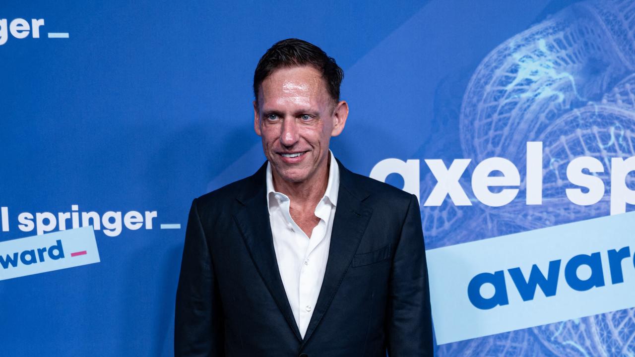 Peter Thiel steht vor einer Wand und lächelt. Auf der Wand ist der Schriftzug "Axel Springer" zu sehen.