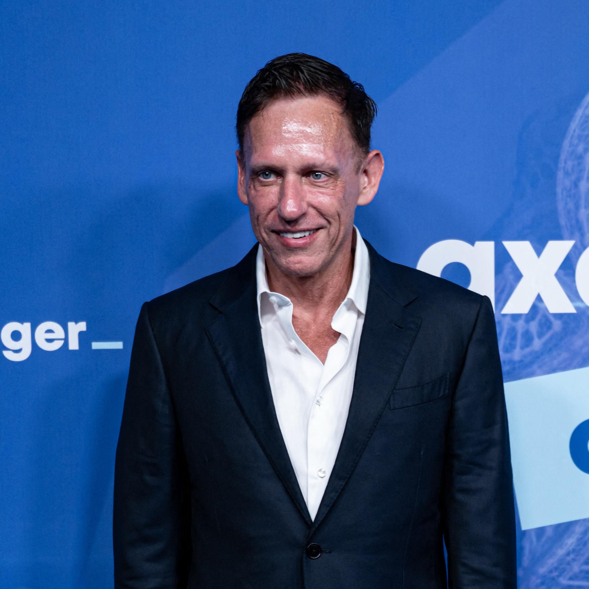 Peter Thiel steht vor einer Wand und lächelt. Auf der Wand ist der Schriftzug "Axel Springer" zu sehen.