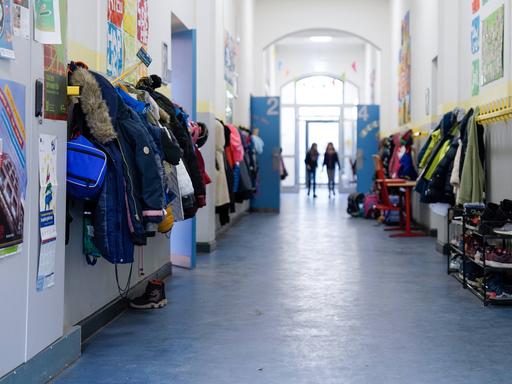 Langer Gang in einer Schule, an der Seite hängen Jacken an der Wand, in der Ferne sind verschwommen zwei Kinder zu sehen.