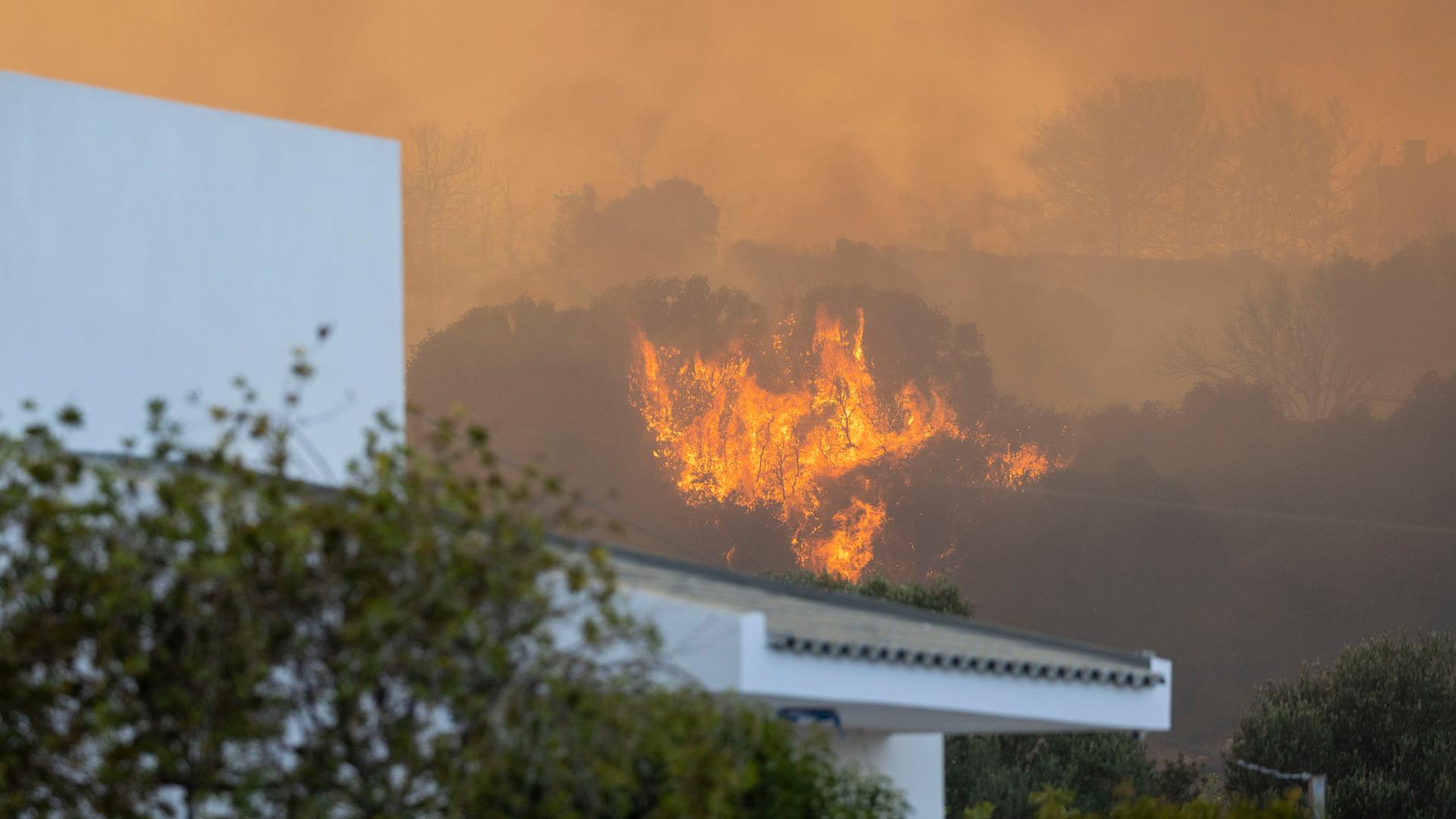 Im Vordergrund ist das Dach eines Ferienhauses zu sehen. Im Hintergrund wütet ein Flammenmeer auf einem Berg auf der griechischen Insel Rhodos. 