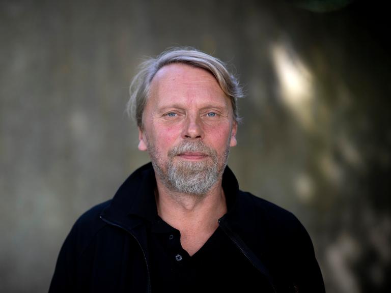 Der Schriftsteller und Journalist Jens Balzer im Portrait mit grauem Bart vor dunklem Hintergrund. 