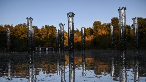 Mahnmal an den "Holocaust mit Kugeln" in der Gedenkstätte Babyn Jar. Unterschiedlich hohe teils silberfarbene Stelen ragen aus dem Wasser. Der Himmel im Hintergrund ist blau.
