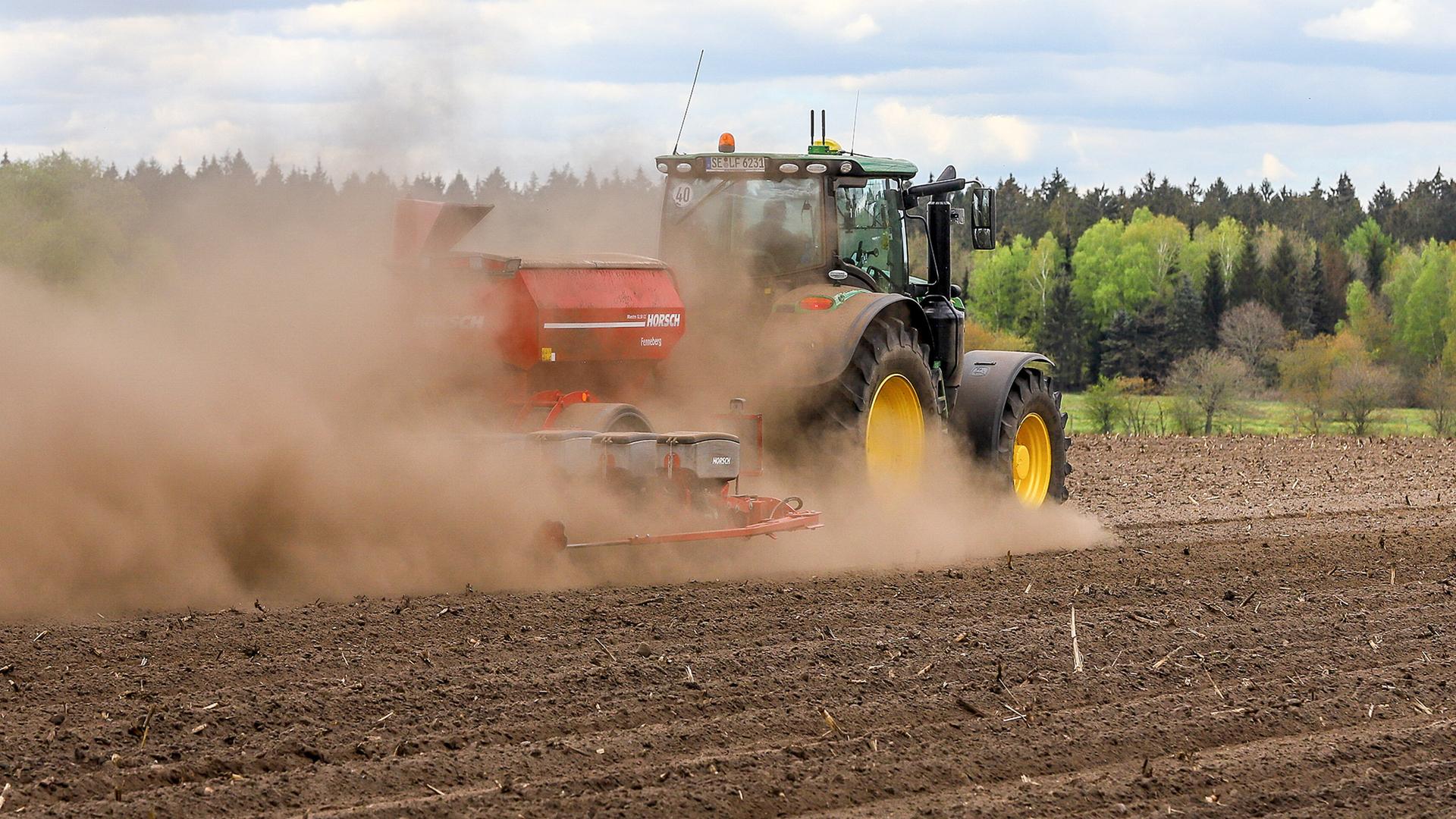Ein Landwirt in einem Traktor fährt über einen trockenen kahlen Acker. Hinter dem Traktor entsteht eine große Staubwolke.
