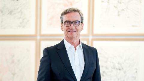 Ein Mann mit grau meliertem Haar und Brille steht in einer Galerie. Er trägt einen schwarzen Anzug über weißem Hemd und lächelt in die Kamera.