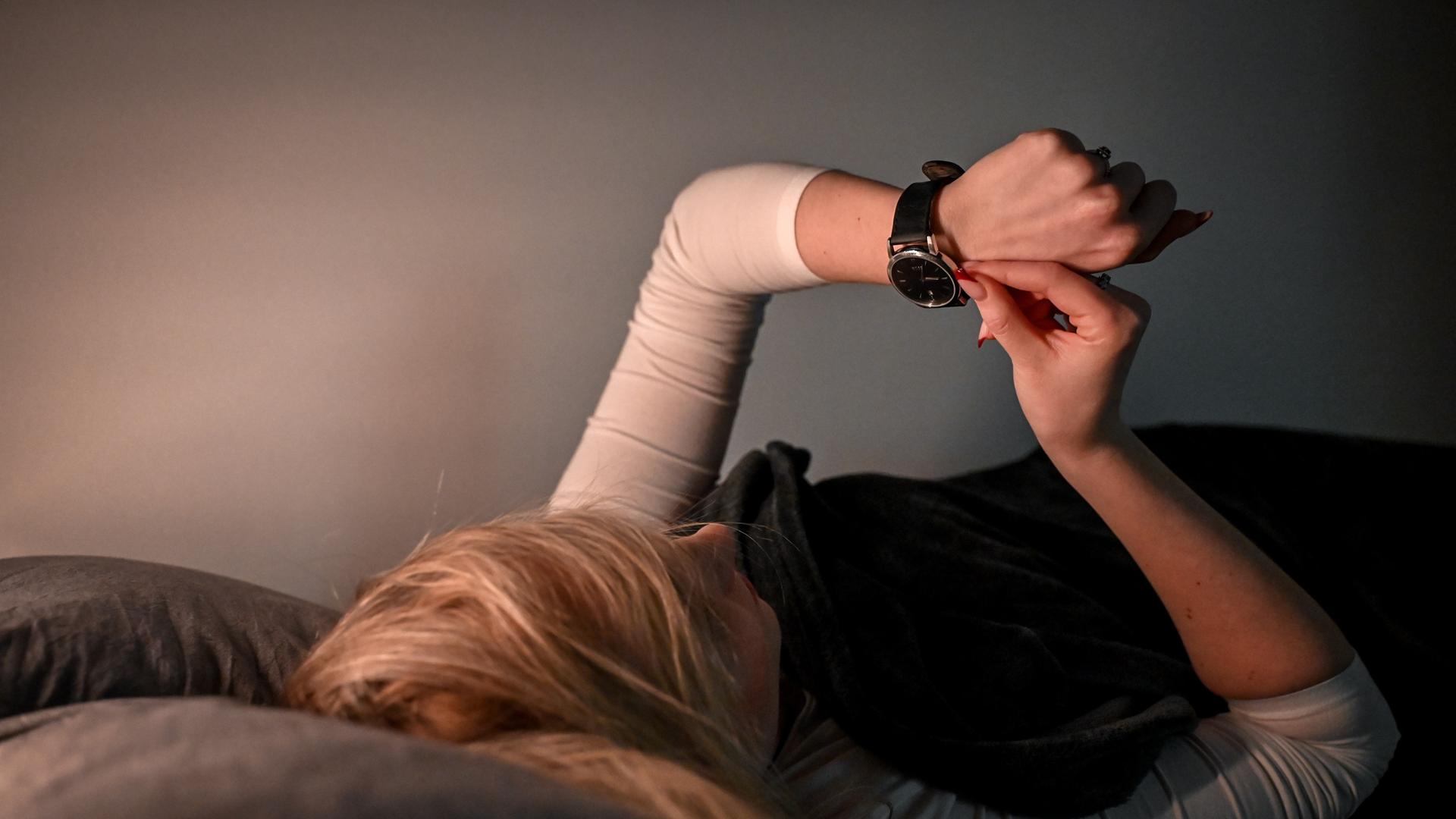 Das Bild zeigt eine Frau im Bett, die auf ihre Armbanduhr schaut und daran dreht.
